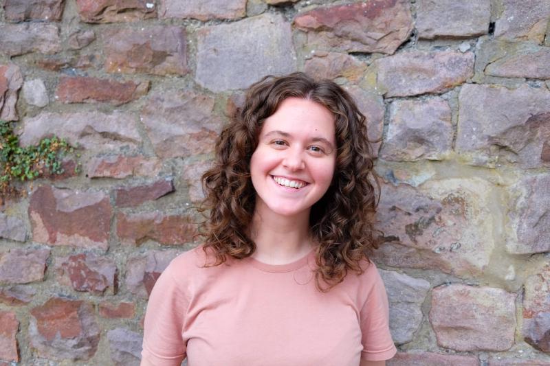 Rae Emily George by a stone wall, she's got curly hair and a big smile