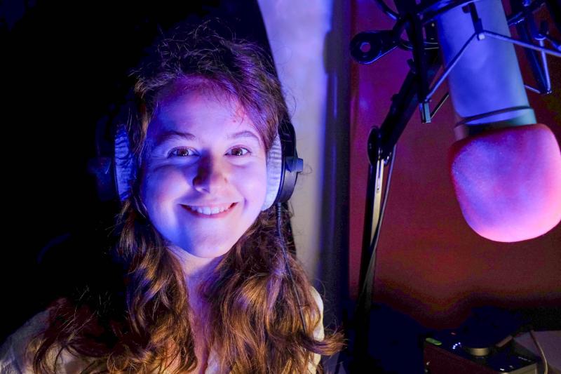 Rae Emily George in a voiceover studio in front of a microphone, smiling at us