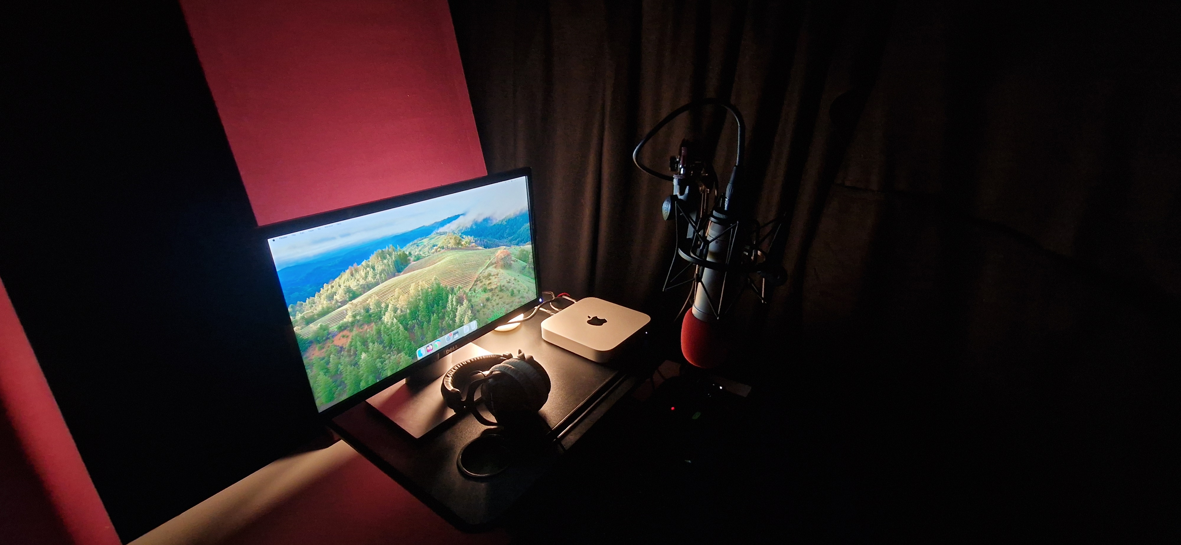 A dark, cozy recording studio surrounded by acoustic curtains and panels. On the desk is my Mac Mini, Screen, RODE NT1-A Microphone, and DT770 Pro Headphones.